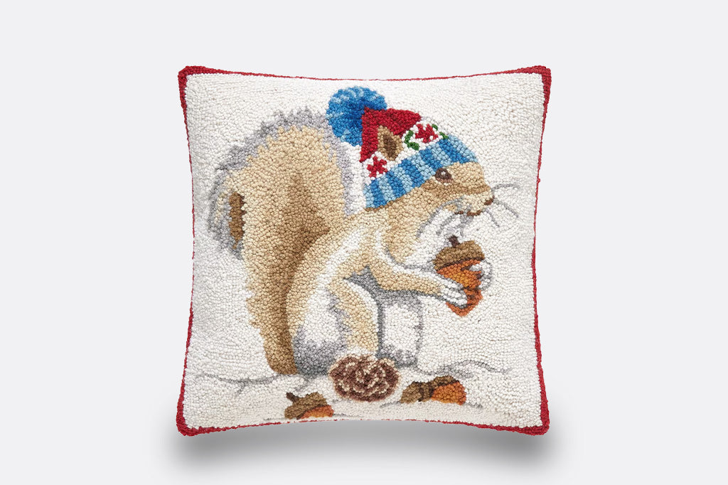 A square decorative throw pillow featuring a tufted embroidered design of a squirrel wearing a blue and red knit hat and holding an acorn, with a red border on a white background.