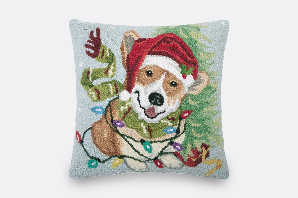 square decorate pillow with a festive corgi wearing a santa hat and winter scarf, entangled in christmas lights in front of a christmas tree with a light blue background pictured on light grey background