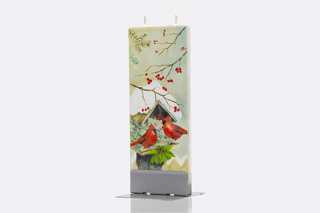 A tall rectangular holiday candle with a printed winter scene featuring two red cardinals perched on a birdhouse among branches with red berries, displayed upright in a gray holder against a light gray background.
