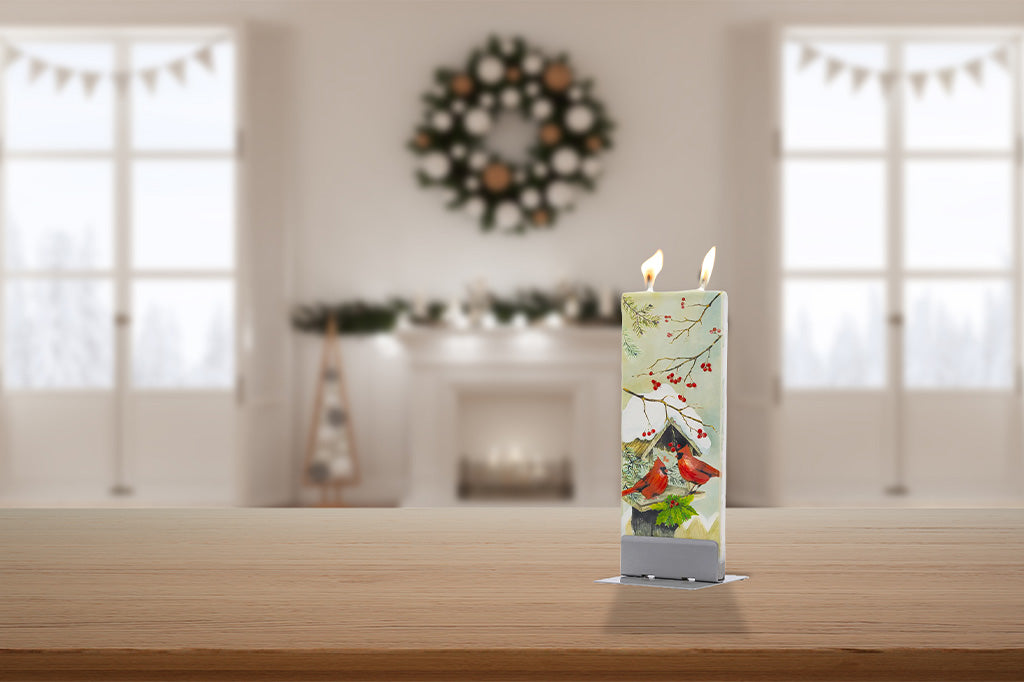 A tall rectangular candle with a winter bird illustration stands lit on a wooden table in a softly blurred living room setting with a fireplace and wreath in the background.