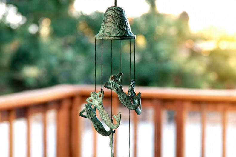 Brass bell wind art with green patina shows reclined mermaid pendants cascading down and moving in the wind on a backyard porch