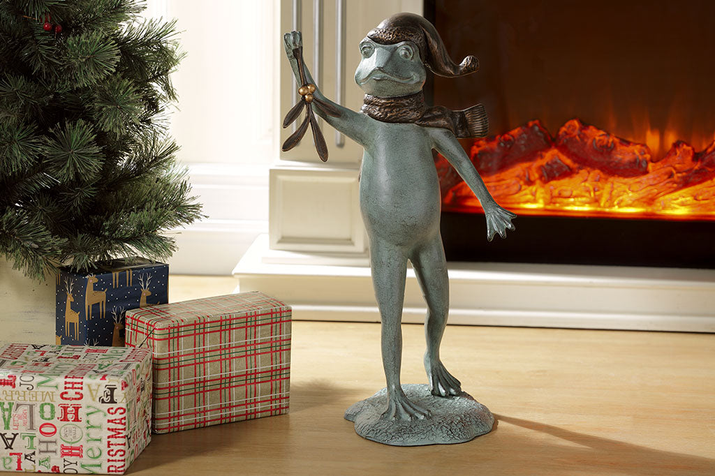 garden sculpture of frog in winter garb holding mistletoe from outstretched webbed hand. It is indoors in front of fireplace and Christmas tree