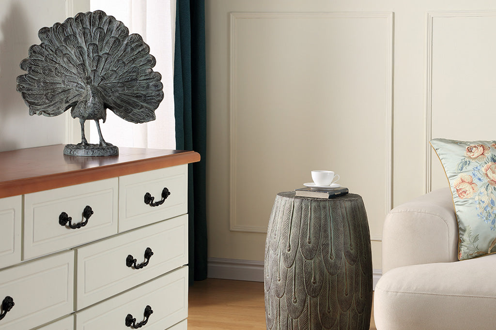 cast metal peacock sculpture shown in room on a dresser with a feather motif garden stool, couch, and teacup nearby