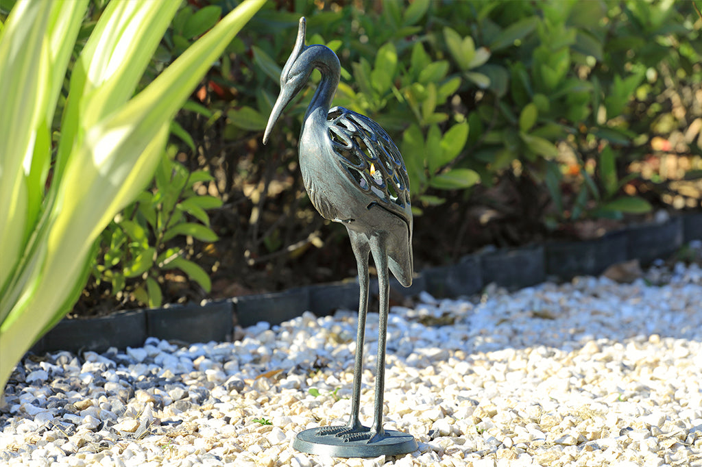 Modern metal heron candleholder with white lit candle face left placed in outdoor garden space 