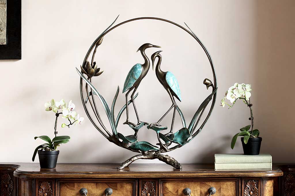 Crane Romantic sculpture shown upon a dresser and surrounded by potted orchids