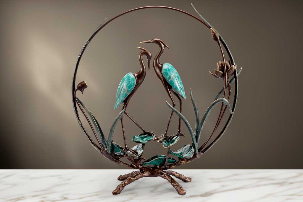 beautifully finished with verdi greens and dark bronze colors, a pair of cranes stands upon sculpted lilies and are enclosed by a hoop with sculpted plants