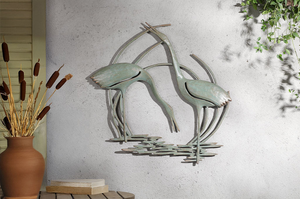 Wall plaque of two cranes standing in reeds and water - hung on concrete outdoor wall by cafe table and hanging plant