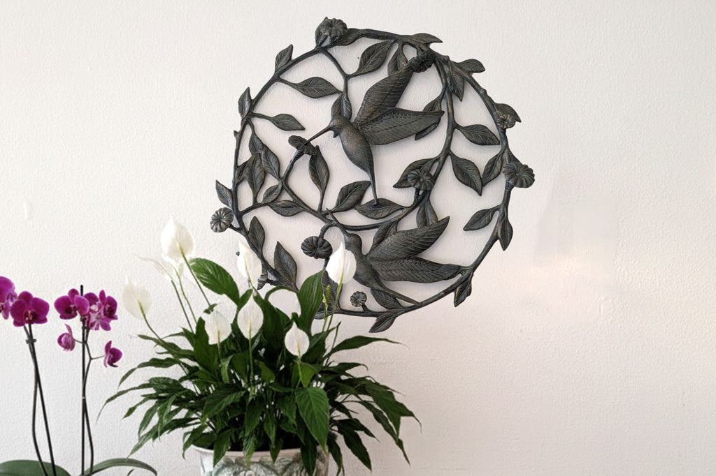 Decorative metal wall art  with hummingbird and vine design next to a potted plant on a white wall in lobby