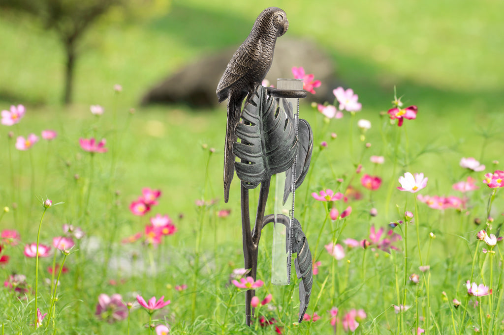 Sculptural rain catcher shaped like a Small parrot sitting on 3 tropical leaves, displayed in a field of wildflowers