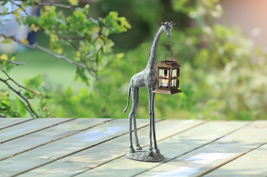 cast metal giraffe holding lantern in mouth sculpture  - safari decor. shown on wood table in backyard setting with oak trees