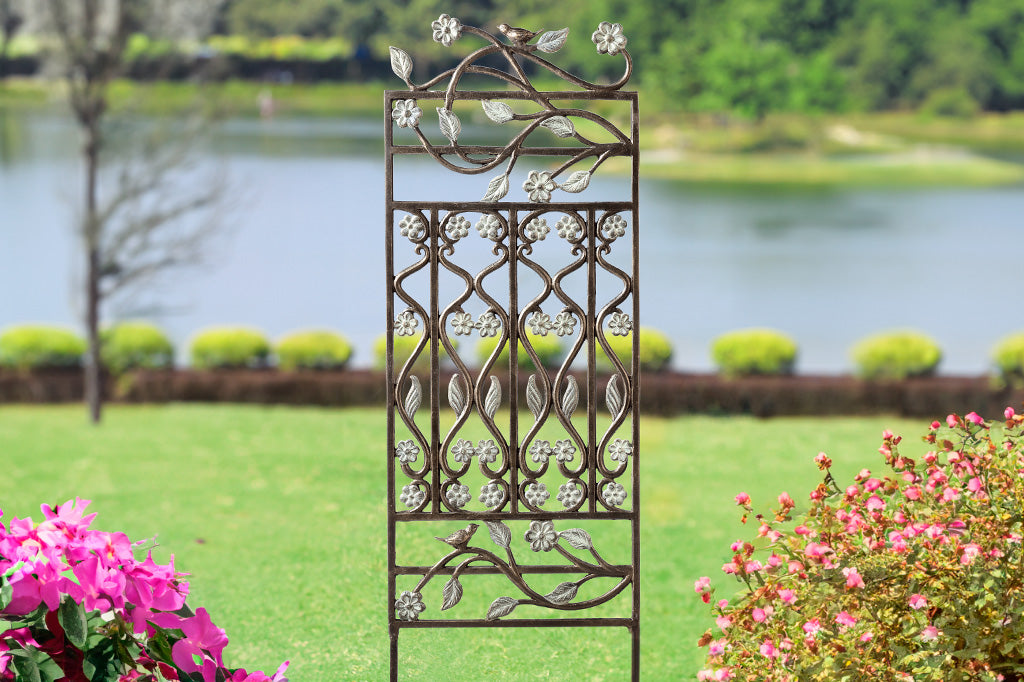 Brindille Trellis features verdi accents, twining branches, and two bird accents. Shown by a lake near blooming bushes