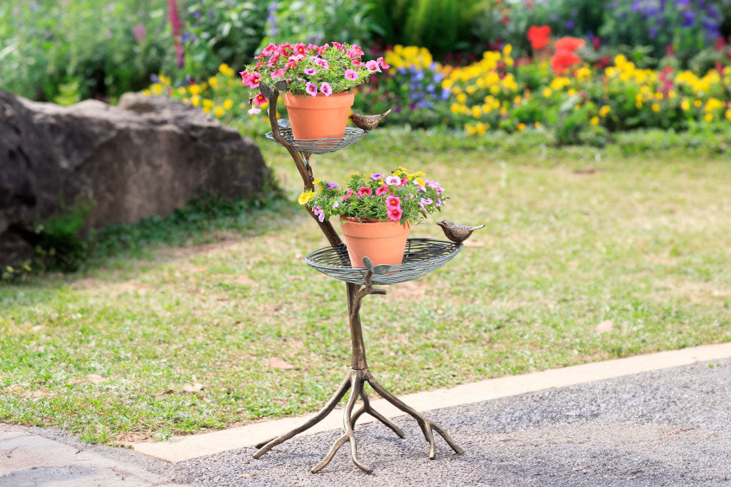 two tier bird feeder/planter shown in a garden on brick path; planter holds potted flowers in terra cots pots, placed in the bird nest of the garden sculpture