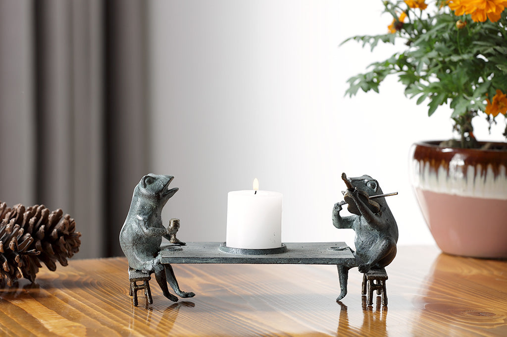 Candleholder sculpture of two frogs at a dining table; one plays violin while the other frog holds goblet. Shown on dining table with pinecones and potted marigolds.