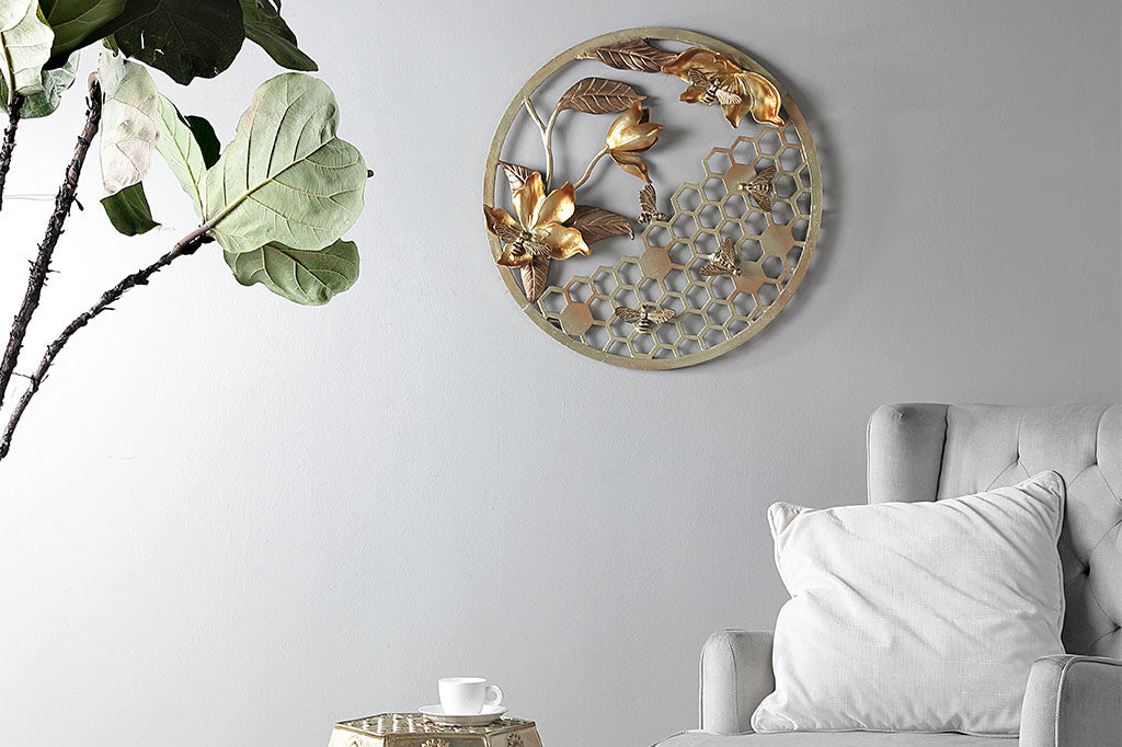 Circular wall plaque in golden cast metal finish featuring bees, honeycomb and blossoms - shown on grey wall near armchair, indoor houseplant, and stool with matching bee motif