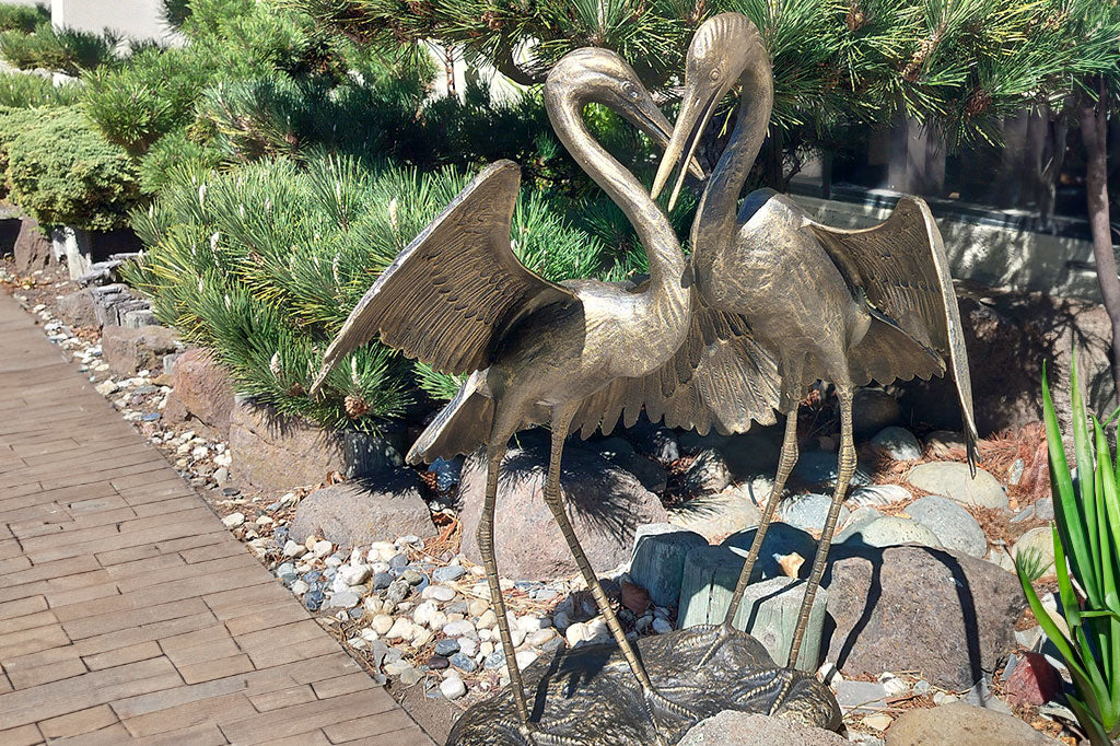 crane sculpture displayed amongst landscaping; nearby is a pine tree and wood-brick path