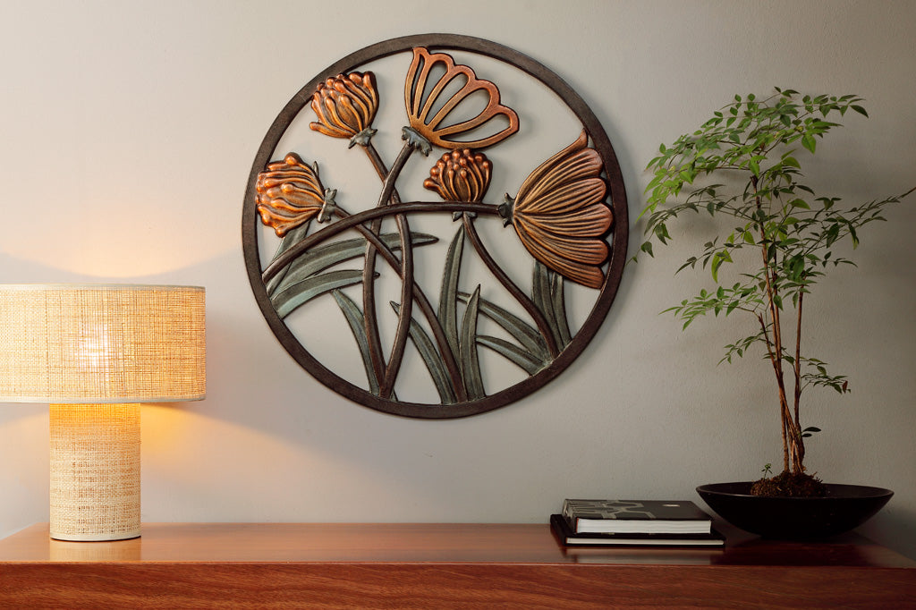 Cast metal circular wall plaque of thistle and blossom shown on console table with potted tree and glowing lamp