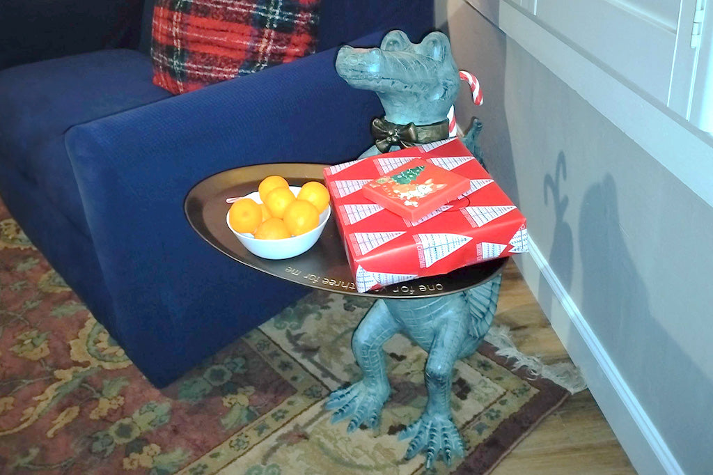 Customer photo of a crocodile butler sculpture in front of a couch; the crocodile wears a bowtie and holds a tray of oranges and wrapped Christmas gifts
