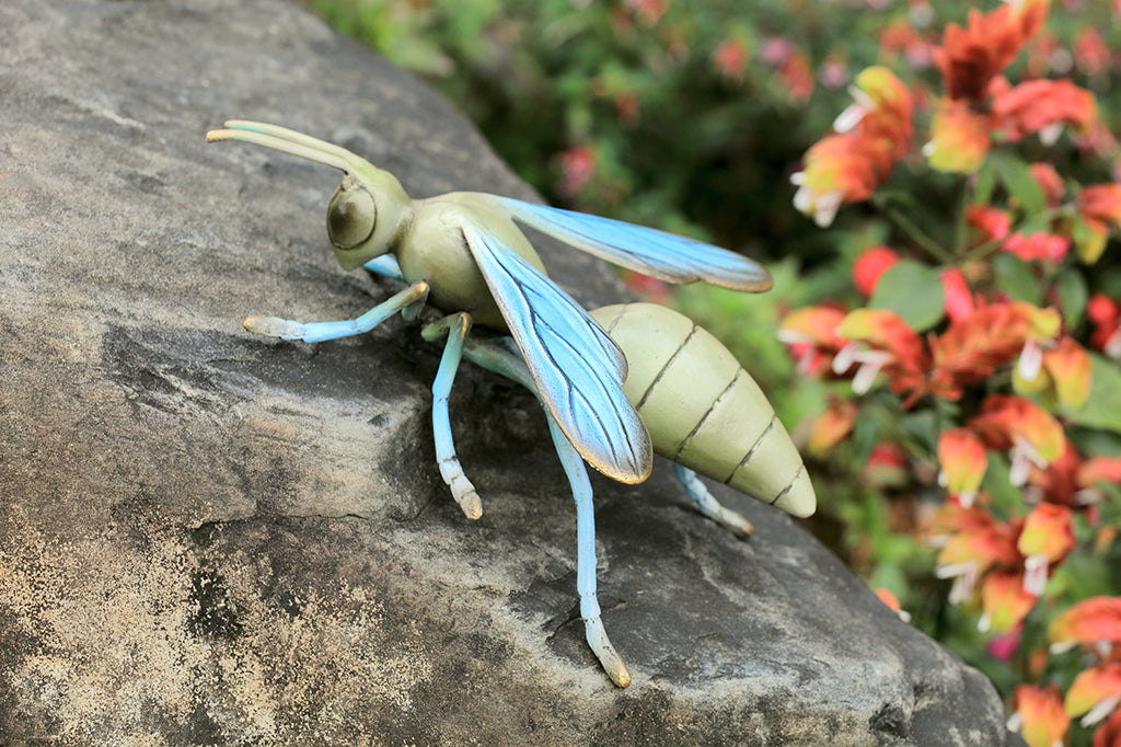 Sculpted metal wasp in soft shades of blue and sage green shown on a rock in a flower garden