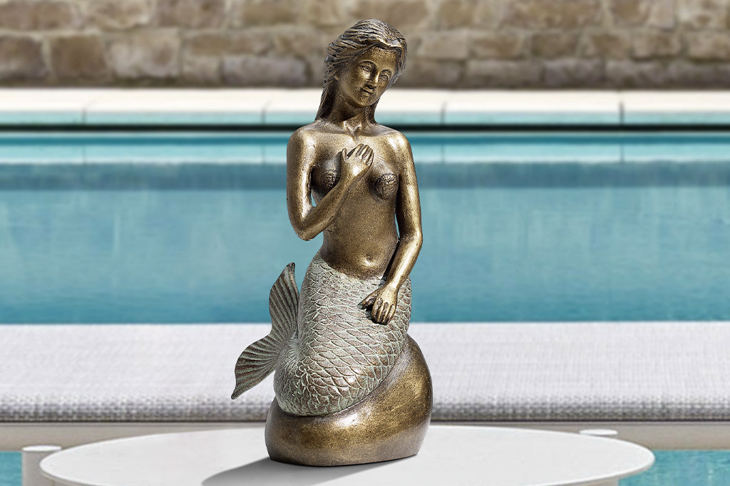 Metal mermaid seated on a rock garden sculpture, staged in pool setting 