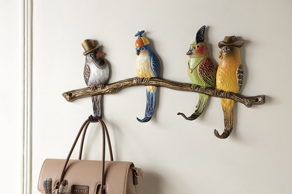 Shown on the wall of a home, 4 sculpted birds on a branch wear stylish headwear; their tailfeathers serve as hooks. First bird hook holds a beige purse by handles