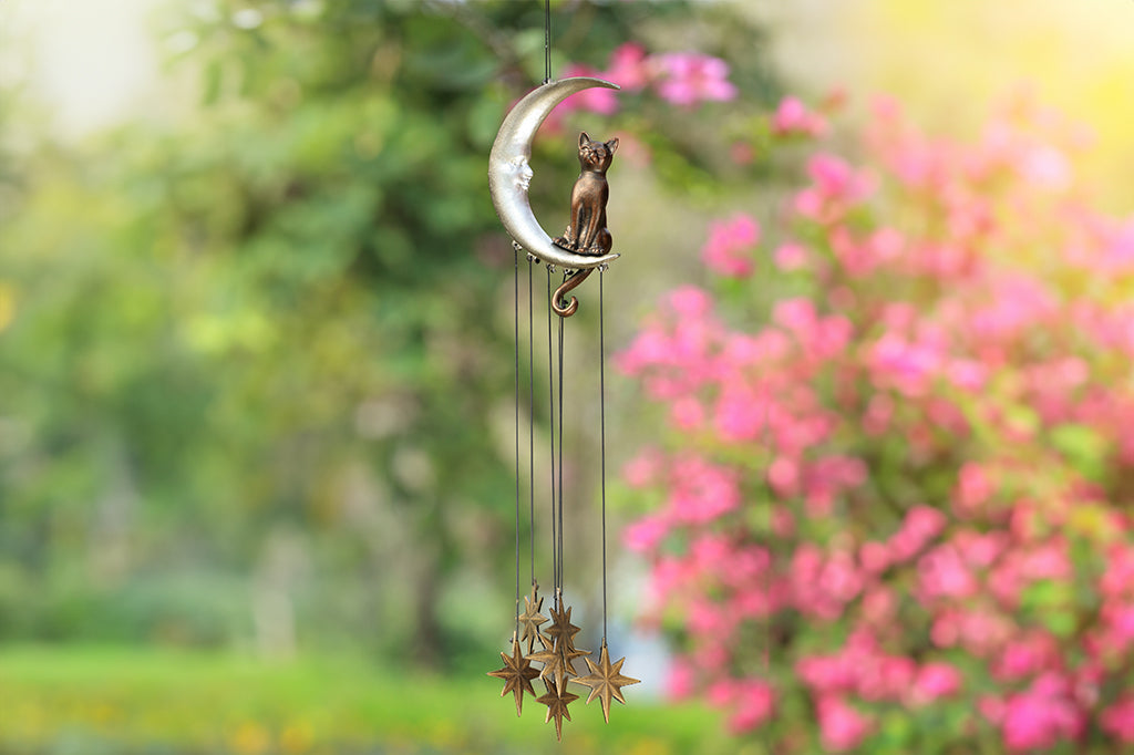 Wind art featuring a cat seated upon a crescent moon with face, 8 point star pendants suspended below. shown in garden with blooming bushes