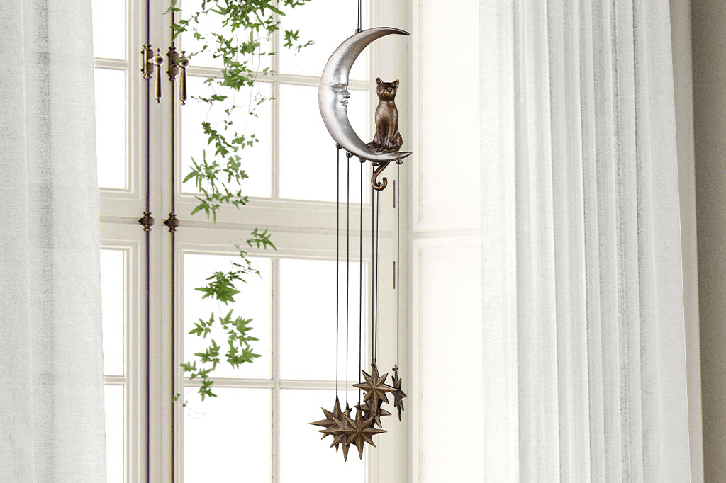 Wind art featuring a cat seated upon a crescent moon with face, 8 point star pendants suspended below. shown in room in front of window