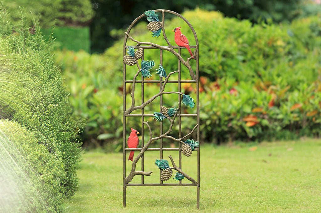 Garden trellis featuring 2 red cardinals, green pine branches, and bronze pine cone accents shown outside in yard with sprinkler spraying on a hedge nearby.