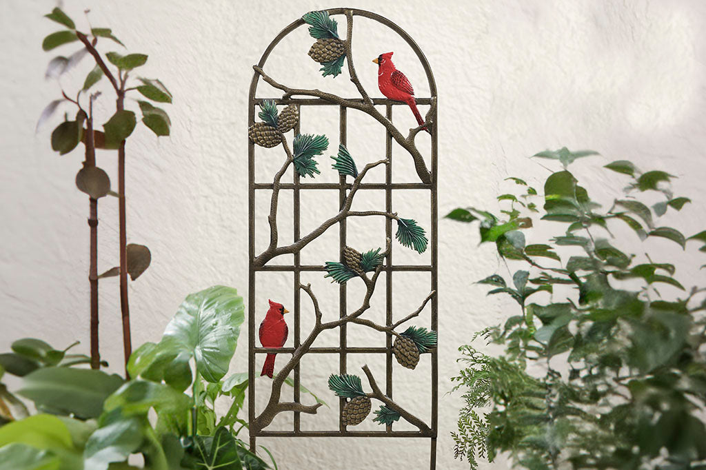 Garden trellis featuring red cardinals, pine branches, and pine cones shown outside of white stucco house amidst foliage. 