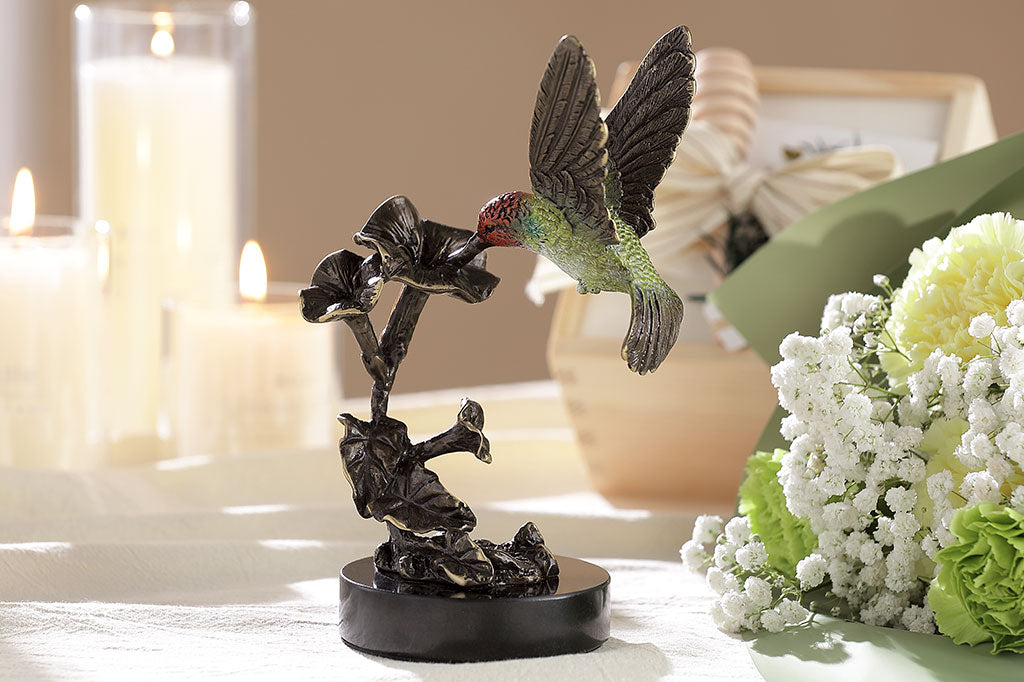 Brass hummingbird sipping from trumpet flower indoor sculpture; shown on table near candleand floral dispay. hummingbird is finished in pale green with red head and bronze wings