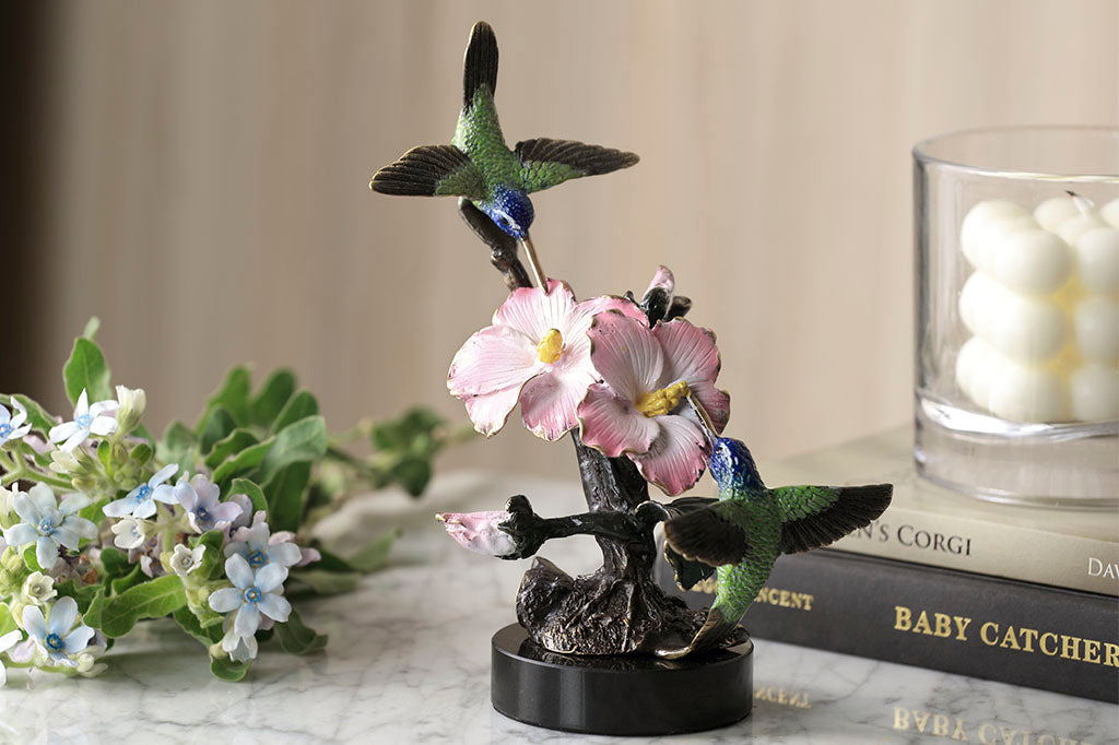 Brass hummingbird sculpture with pink blossoms, indoor sculpture shown on table with books, candle, and blossoms