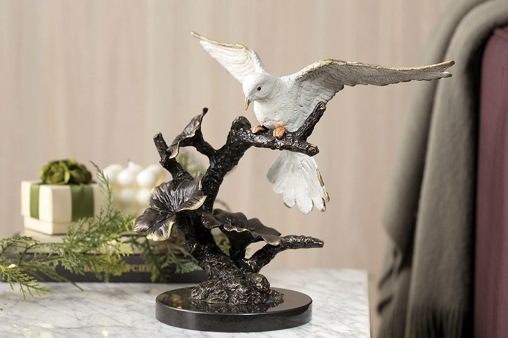 Brass sculpture of dove, with outstretched wings on a flowering branch shown indoors on side table with gifts in background