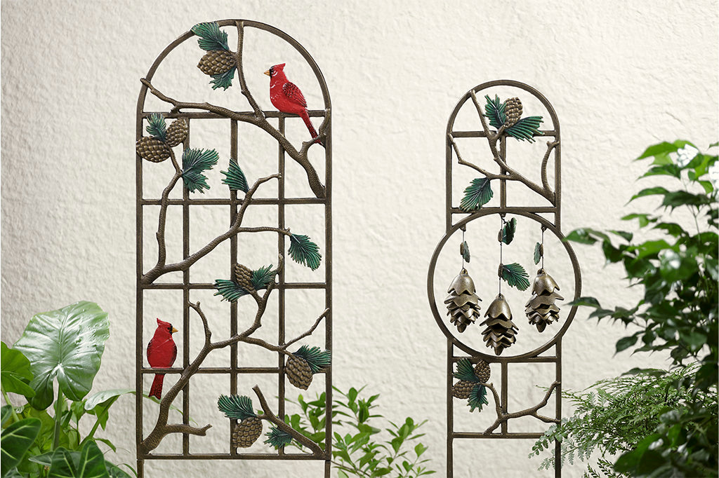 Two garden trellises featuring pine branches and pine cones shown outside of white stucco house. Trellis on left is taller and has 2 red cardinals; trellis on right has hoop focal point and contains  hanging pinecone shaped pendants