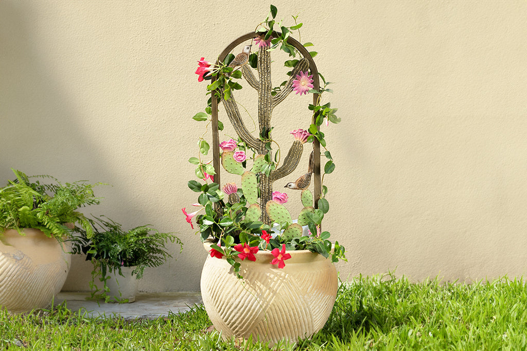 garden trellis shown in flower pot in a yard with grass and stucco building in background. Pink blooming passionflower draped upon it; it features two cacti with pink blooms and cast metal birds