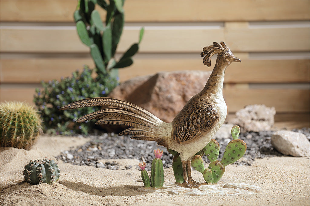 A statue of a roadrunner stood between a group of cacti placed in a sand and rock filled garden