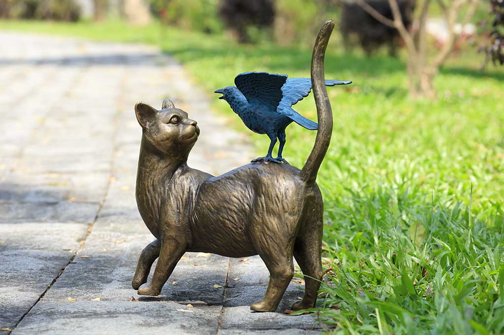 Bronze sculpture of a cat with a bird on its back in an outdoor setting