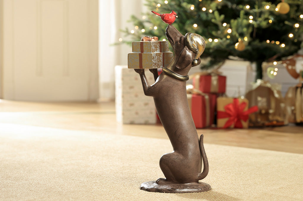 Shown in living room with christmas tree in the background, the Hello, Friend Garden Sculpture features a Dachshund sculpture holding wrapped gifts while a red cardinal perches on its nose.