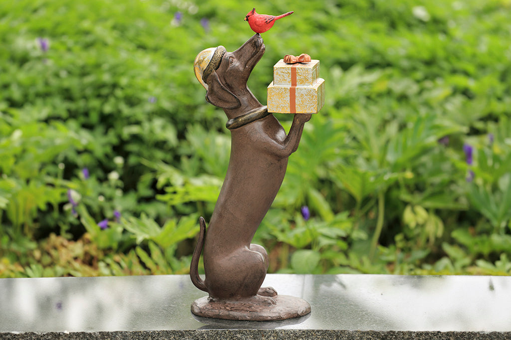 Shown on marble bench in garden: Hello, Friend Garden Sculpture features a Dachshund sculpture holding wrapped gifts while a red cardinal perches on its nose.