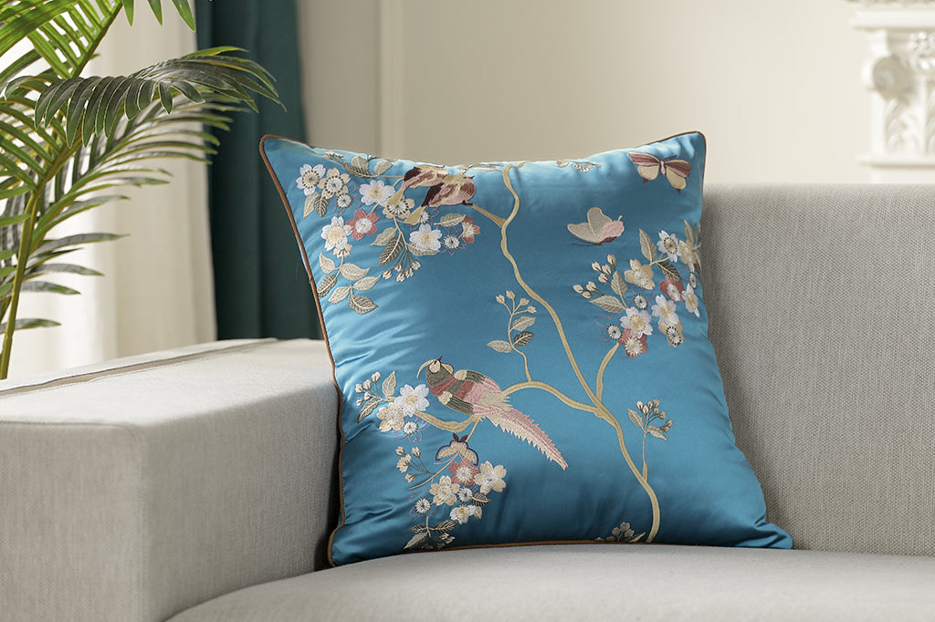 dark blue satin pillow with embroidered flowers, birds, branches, and fluttering butterflies placed on couch 