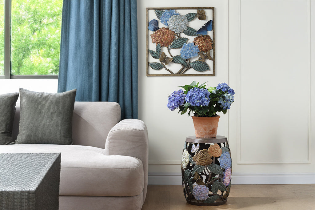 Wall plaque features hydrangea blossoms, butterflies and green leaves. Shown in living room with potted hydrangeas and blue curtains and hydrangea stool VB35442