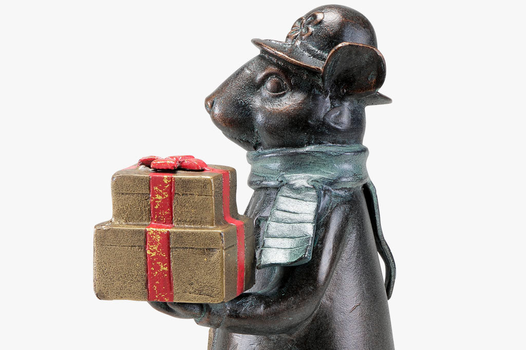 Close up view of Christmas metal sculpture: Mouse dressed in wintercoat with purse holds 2 gold and red wrapped gifts in paw; she wears a green scarf and a hat with bow on it.