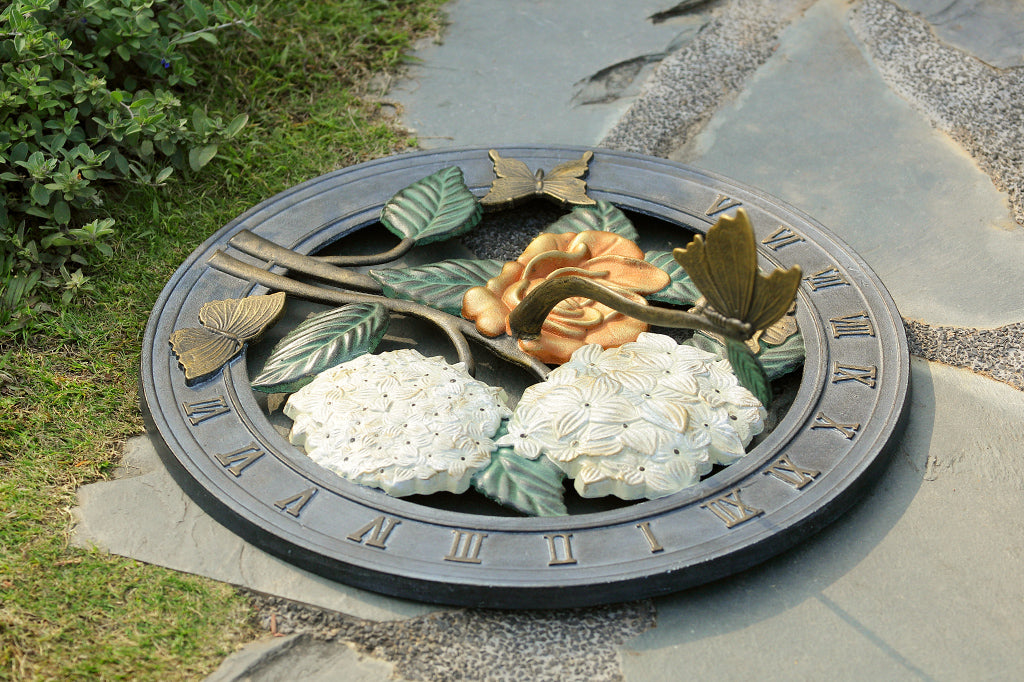 Sundial in cast metal with roman numerals, and a butterfly on the shadow-casting gnomon. Features butterflies, hydrangea, and roses. shown on garden path