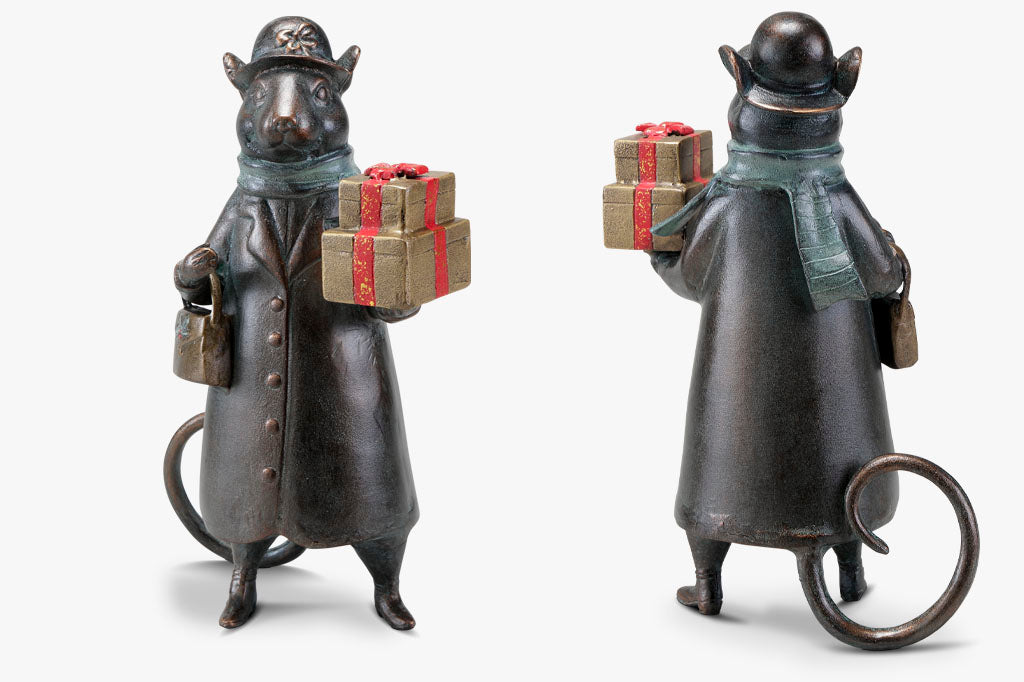 Front and back view of a mouse sculpture. Mouse lady is dressed in wintercoat, holds a purse and 2 gold and red wrapped gifts in paw; she wears a green scarf and a hat with bow on it. She has a curled tail behind her.