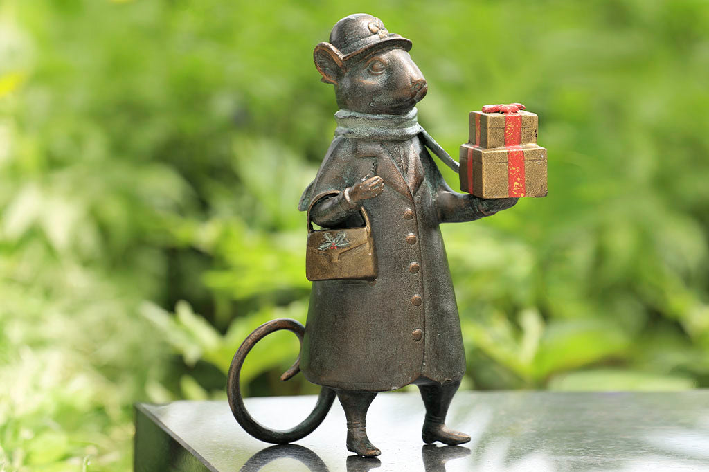 Holiday metal sculpture shown on marble bench in leafy garden. Sculpture shows mouse dressed in wintercoat with purse holds 2 gold and red wrapped gifts in paw; she wears a green scarf and a hat with bow on it.