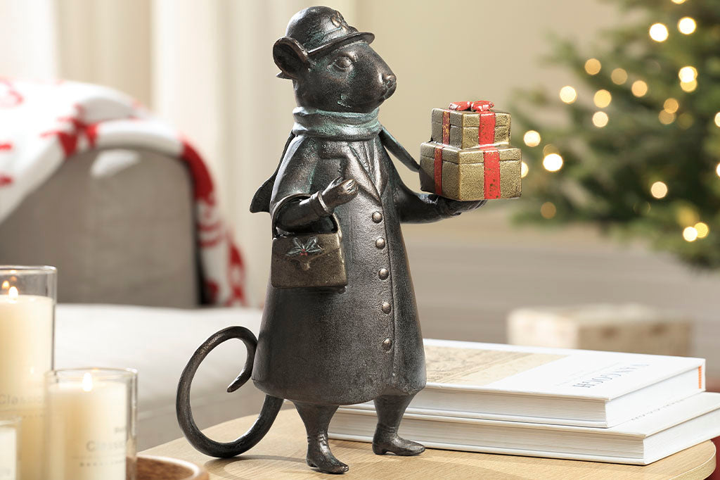 Holiday metal sculpture shown in Christmas scene on table near tree with holiday lights. Mouse dressed in wintercoat with purse holds 2 gold and red wrapped gifts in paw; she wears a green scarf and a hat with bow on it.