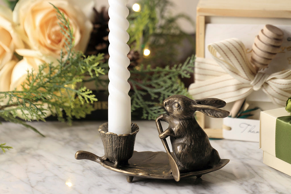 taper candleholder in brass; rabbit rowing a leaf with a blossom-shaped candleholder cup - holding white candle in scene with fresh roses and wrapped gifts