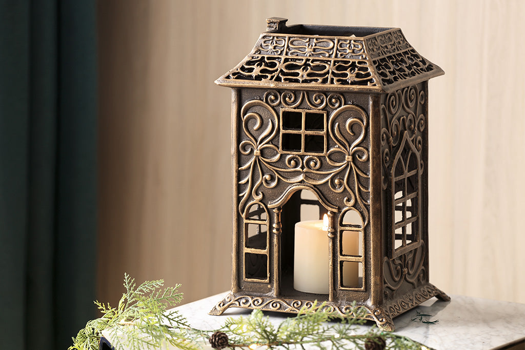 Cast metal house candleholder styled with Victorian style "gingerbread" embellishments like scrollwork and arched window fretwork, shown on countertop with faux evergreens