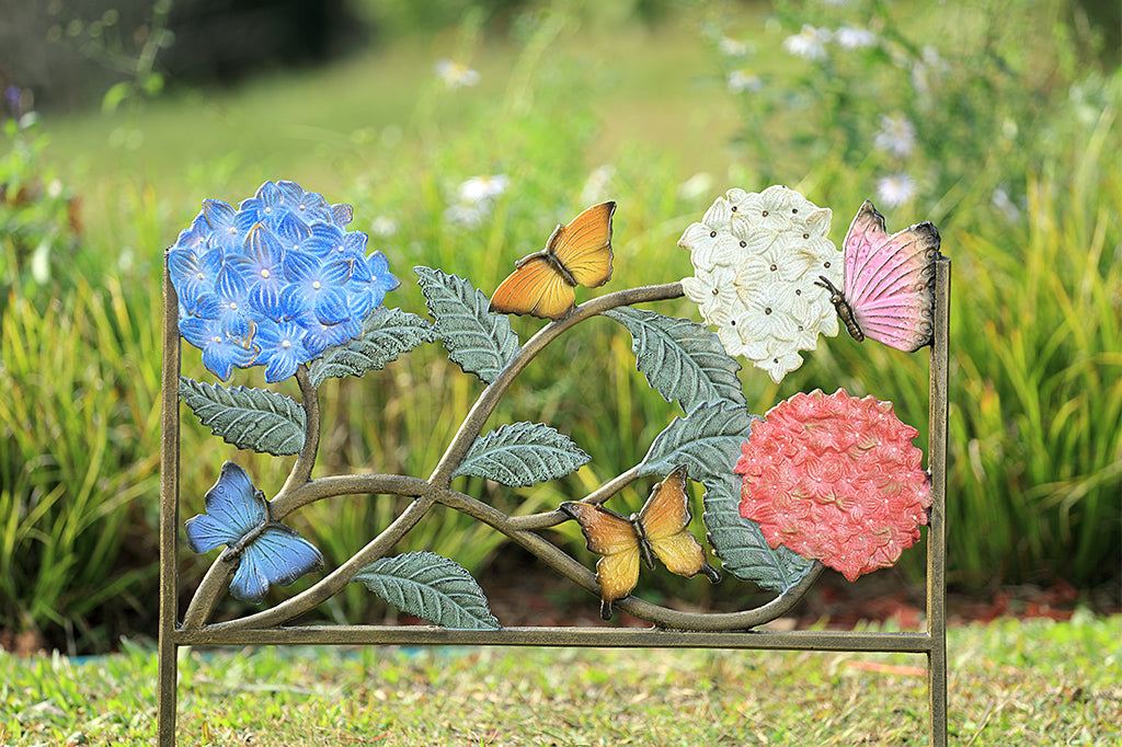 4 butterflies scatter amongst bold hydrangea blossoms of blue, white, and pink with green leaves in a garden edging frame.