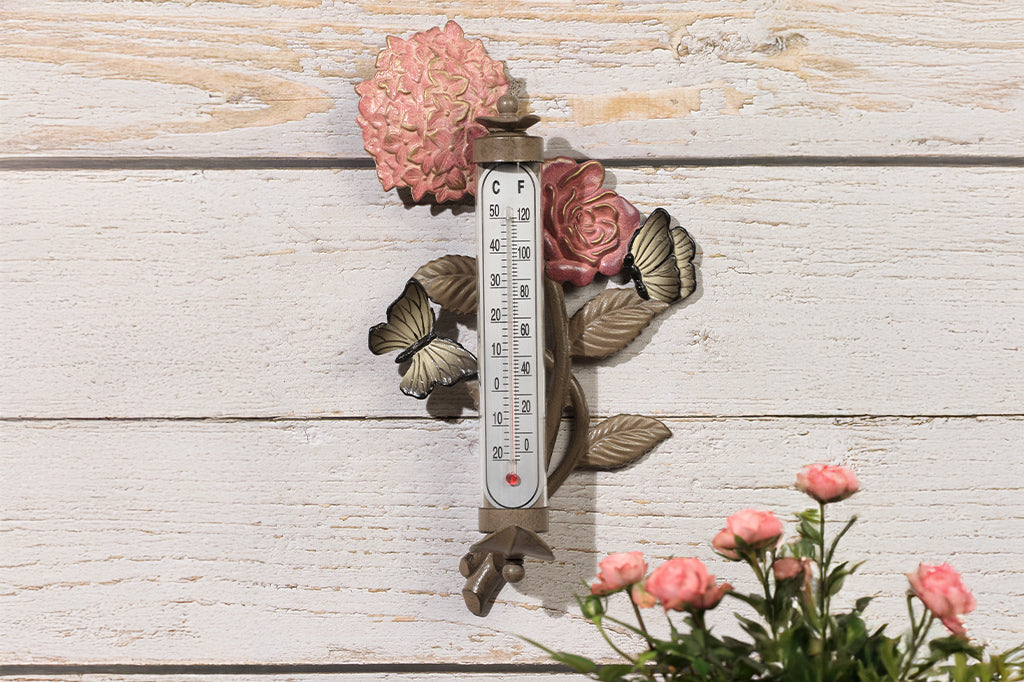 Wall mounted thermometer with cast metal accents of pink rose and hydrangea blossoms, butterflies, and leaves, shown on wood wall by planting of tea roses