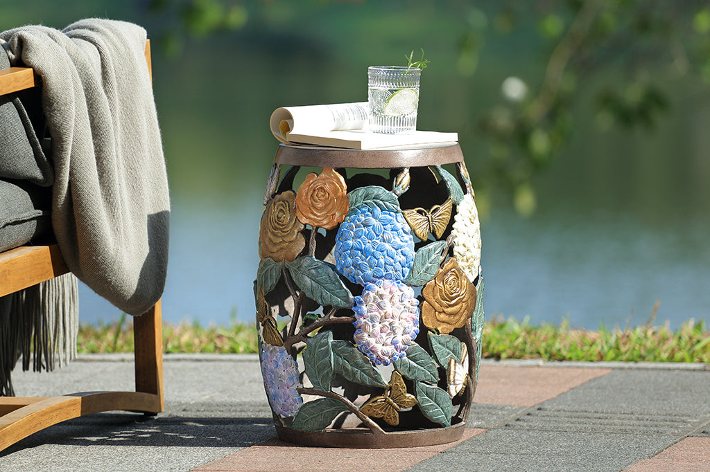 Cast metal garden stool features beautiful roses, butterflies, and the vibrant hues of hydrangeas. Solid top shown with book and glass of water - shown outside by a lake and chair with throw blanket.