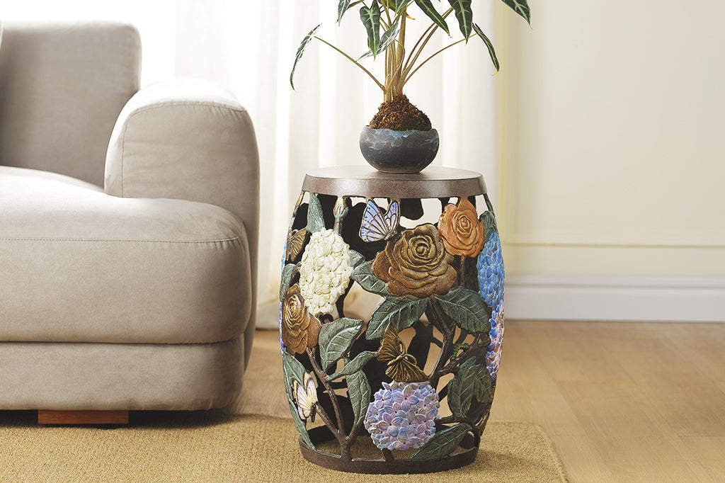 Cast metal garden stool features beautiful roses, butterflies, and the vibrant hues of hydrangeas. Solid top shown with potted plant inside a living room scene.